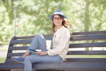 A happy young student with a tablet pc and a disposable coffee cup sitting on the bench in a summer park.