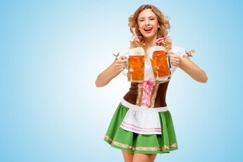 Young sexy Oktoberfest woman wearing a traditional Bavarian dress dirndl serving two beer mugs with happy smile on blue background.
