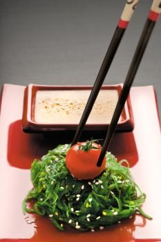 A close-up of chopsticks holding a red-ripe tomato on chuka salad along with gomadare sauce in the background.