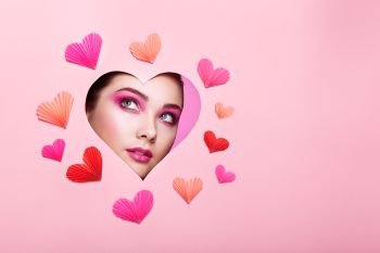 Conceptual photo of Valentine’s day. Face of Girl with Festive Pink Makeup. Paper hearts on a pink background. Love symbols Valentines day
