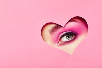 Conceptual photo of Valentine’s day. Eye of Girl with Festive Pink Makeup. Paper heart on a pink background. Love symbols Valentines day