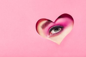 Conceptual photo of Valentine’s day. Eye of Girl with Festive Pink Makeup. Paper heart on a pink background. Love symbols Valentines day
