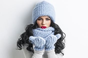 Woman in winter clothes blowing on palms. Beautiful woman in blue winter hat, scarf and mittens blowing on palms