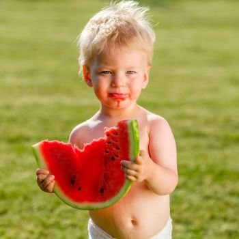 Portrait of toddler child outdoors. Rural scene with one year old baby boy eating watermelon slice in the garden. Dirty messy face of happy kid.
