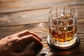 Glass of whiskey with ice cubes and man’s hand on rustic wooden table with copy-space. Alcoholism concept.