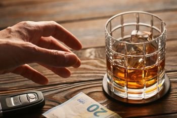 Man’s hand reaching to glass of whiskey or alcohol drink with ice cubes and car key on rustic wooden table. Drink and drive and alcoholism concept. Safe and responsible driving concept.