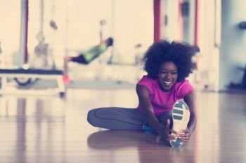 happy young african american woman in a gym stretching and warming up before workout young mab exercising with dumbbells in background. woman in a gym stretching and warming up man in background worki