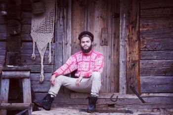 portrait of young hipster,  man with beard in front of old vintage wooden house. portrait of young hipster in front of wooden house