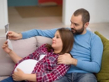 pregnant couple looking baby’s ultrasound. Young pregnant couple looking baby’s ultrasound photo while relaxing on sofa at home