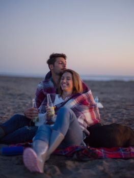 Young Couple Relaxing By The Fire, Drinking A Beer Or A Drink From The Bottle.. Loving Young Couple Sitting On The Beach beside Campfire drinkin