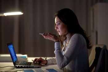 business, technology, overwork, deadline and people concept - woman with laptop and smartphone using voice command recorder at night office. woman using voice command recorder on smartphone
