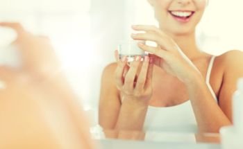 beauty, skin care and people concept - close up of smiling young woman with face cream mirror reflection at home bathroom. happy woman with face cream at bathroom