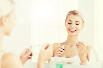 beauty, make up, cosmetics, morning and people concept - smiling young woman with lipstick and applicator applying makeup and looking to mirror at home bathroom. woman with lipstick applying make up at bathroom