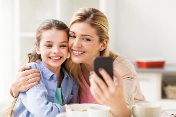 family, technology and people concept - happy mother and daughter with smartphone having breakfast and taking selfie at home kitchen. happy family taking selfie by smartphone at home