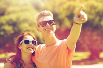 holidays, vacation, love and people concept - happy smiling teenage couple hugging and pointing finger to something at summer park. happy couple pointing finger at summer park