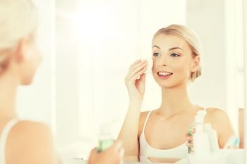 beauty, skin care and people concept - smiling young woman applying lotion to cotton disc for washing her face at bathroom. young woman with lotion washing face at bathroom