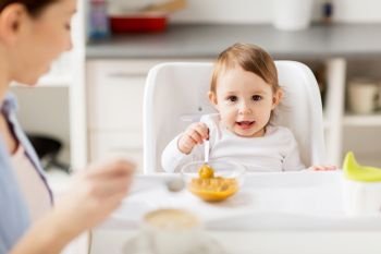 family, food, breakfast and people concept - happy mother with coffee cup and little baby sitting in highchair and eating puree at home kitchen. happy mother and baby having breakfast at home