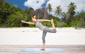 fitness, sport and people concept - woman doing yoga in lord of the dance pose on mat over exotic tropical beach background. woman in yoga lord of the dance pose on beach