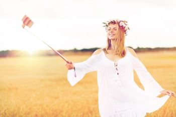 technology, summer holidays, vacation and people concept - smiling young woman in wreath of flowers taking picture by smartphone selfie stick on cereal field. happy young woman taking selfie by smartphone