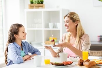 family, holidays and people concept - happy girl and mother holding piece of birthday cake with burning candle at home kitchen. happy family with birthday cake at home kitchen