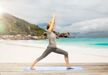 fitness, people and healthy lifestyle concept - happy woman doing yoga warrior pose on mat over exotic tropical beach background. happy woman doing yoga warrior pose on beach