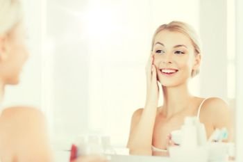 beauty, skin care and people concept - smiling young woman applying cream to face and looking to mirror at home bathroom. happy woman applying cream to face at bathroom