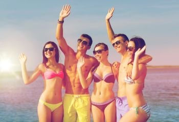 friendship, sea, holidays, gesture and people concept - group of smiling friends wearing swimwear and sunglasses waving hands on beach. smiling friends in sunglasses on summer beach