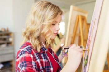 art school, creativity and people concept - student girl or artist with easel, earphones and paint brush painting at studio. student girl with easel painting at art school
