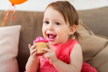 childhood, people and celebration concept - happy baby girl eating cupcake on birthday party at home. happy baby girl eating cupcake on birthday party