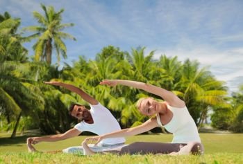 fitness, sport and people concept - couple making yoga exercises outdoors over exotic natural background with palm trees. happy couple making yoga exercises outdoors