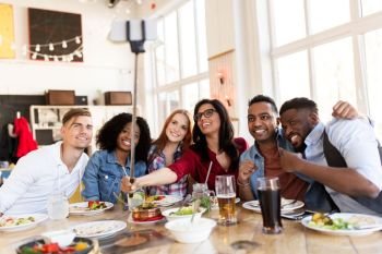 leisure, technology and people concept - group of happy international friends eating and taking picture by smartphone selfie stick at restaurant. friends eating and taking selfie at restaurant