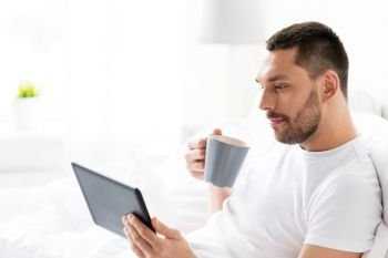 technology, internet, communication and people concept - young man with tablet pc computer drinking coffee in bed at home bedroom. man with tablet pc drinking coffee in bed at home