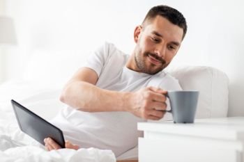 technology, internet, communication and people concept - young man with tablet pc computer drinking coffee in bed at home bedroom. man with tablet pc drinking coffee in bed at home
