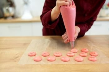 cooking, baking and people concept - chef with confectionery bag squeezing macaron batter or meringue cream to baking paper at pastry shop kitchen. chef with injector squeezing macaron batter