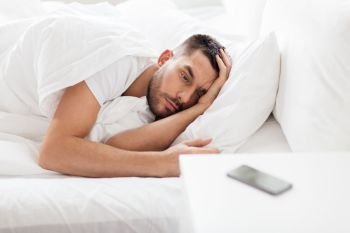 technology, internet, communication and people concept - young man lying in bed at home in morning and looking at smartphone. young man looking at smartphone