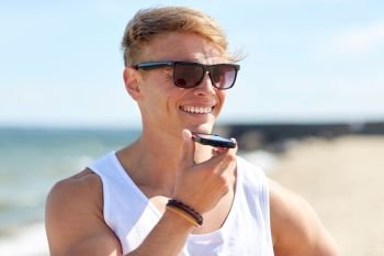 summer holidays and people concept - happy smiling young man in sunglasses using voice command recorder on smartphone on beach. smiling man calling on smartphone on summer beach