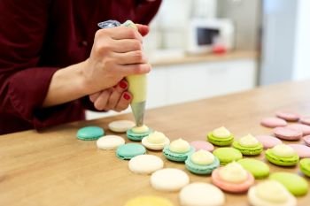 cooking, food and baking concept - chef with confectionery bag squeezing cream filling to macarons shells at pastry shop kitchen. chef with injector squeezing filling to macarons