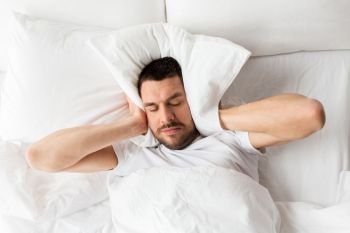 people, bedtime and rest concept - man lying in bed with pillow suffering from noise at home. man in bed with pillow suffering from noise
