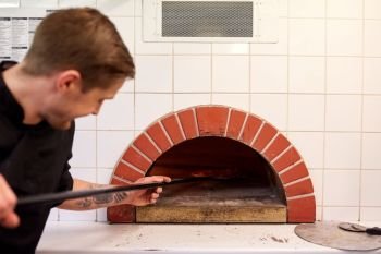 food cooking, culinary and people concept - cook or baker with peel placing pizza to oven at pizzeria. man with peel placing pizza to oven at pizzeria