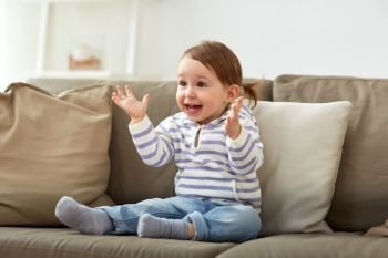 childhood, emotions and people concept - happy smiling baby girl sitting on sofa and clapping hands at home. happy smiling baby girl sitting on sofa at home