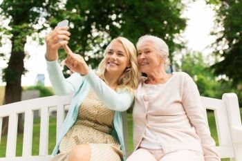 family, technology and people concept - happy smiling young daughter and senior mother with smartphone sitting on park bench and taking selfie. daughter and senior mother taking selfie at park