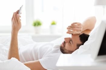 technology, internet, communication and people concept - happy young man texting on smartphone in bed at home in morning. young man with smartphone in bed in morning