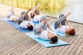 yoga, fitness, sport, and healthy lifestyle concept - group of people making supine pigeon pose on mat outdoors on river or lake berth. people making yoga in supine pigeon pose outdoors