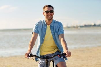 people, leisure and lifestyle concept - happy young man in sunglasses riding bicycle along summer beach. happy man riding bicycle along summer beach