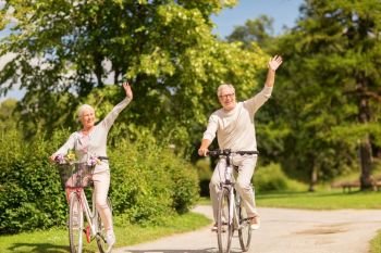 active old age, people and lifestyle concept - happy senior couple riding bicycles and waving hand at summer park. happy senior couple riding bicycles at park