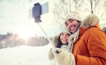 people, season, love, technology and leisure concept - happy couple taking picture with smartphone selfie stick on over winter background. happy couple taking selfie by smartphone in winter