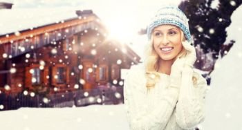 winter, vacation, christmas and people concept - smiling young woman in hat, sweater and gloves over wooden country house and snowflakes background. smiling young woman in winter hat and sweater