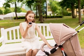 motherhood, technology and people concept - happy mother with baby in stroller using voice command recorder on smartphone at summer park. mother with stroller and smartphone at summer park