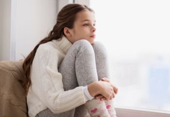 childhood, sadness and people concept - sad beautiful girl in sweater sitting on sill at home window in winter. sad girl sitting on sill at home window in winter