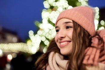 holidays and people concept - portrait of beautiful happy young woman over christmas tree lights in winter evening. happy young woman at christmas market in winter